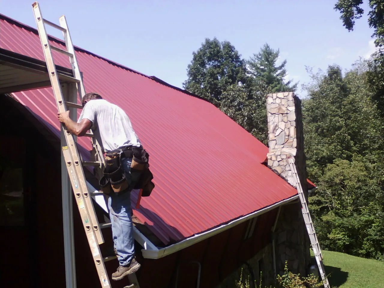 Roofer on a ladder beside a red metal roof and stone chimney — Soffit & Fascia Repair by Roofing Brothers in Mount Holly Springs